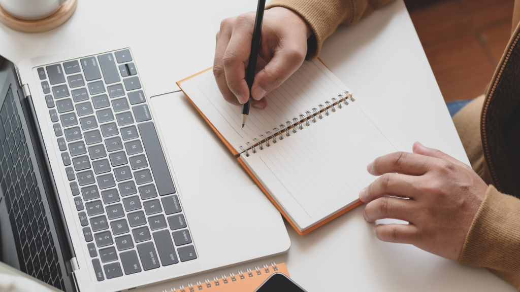 Person taking notes by hand while working on a laptop, representing training and evidence review